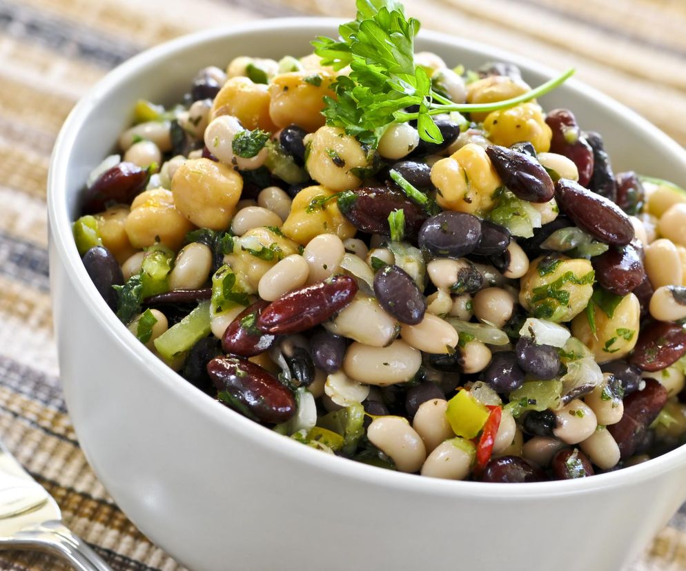 Miska z sałatką ze strączków, zawierająca ciecierzycę, białą fasolę i ciemną fasolę, posypana świeżą pietruszką, na tle beżowego obrusu. Taki zdrowy posiłek pomoże schudnąć i obniżyć cholesterol, o czym przeczytasz więcej na Poradnik Zdrowie.