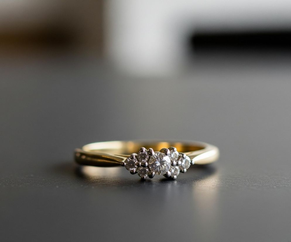 Centralnie na obrazie znajduje się złoty pierścionek z trzema diamentami, umieszczony na ciemnej, gładkiej powierzchni. Dwa mniejsze diamenty flankują większy, centralny kamień, wszystkie osadzone w białym metalu. Pierścionek odbija subtelne światło, tworząc delikatne refleksy na powierzchni stołu. Tło jest rozmyte, tworząc gradient od jasnego, mlecznego odcienia w górnej części do ciemniejszego brązu i szarości w pozostałych obszarach.