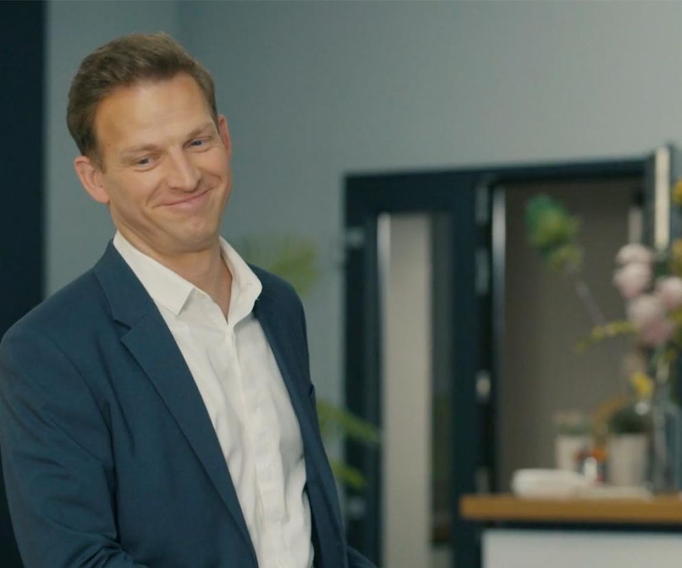 Michał Rolnicki jako Łukasz Sadowski z serialu „Barwy szczęścia uśmiecha się na zbliżeniu w niebieskiej marynarce i białej koszuli, z lekko uniesionymi brwiami, patrząc w bok. Artykuły o jego losach w „Barwach szczęścia” przeczytasz na stronie Super Seriale.