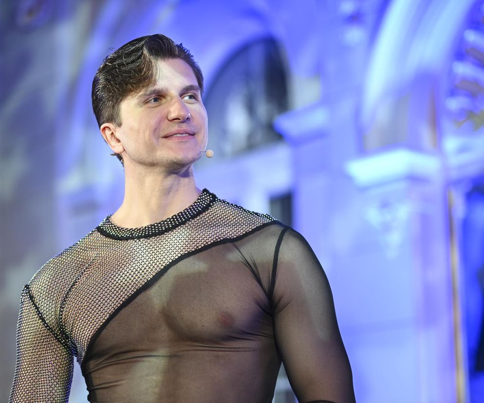 Uśmiechnięty Michał Kassin w błyszczącej, czarnej, prześwitującej siateczkowej koszulce z ozdobnym dekoltem, na tle niebieskiego oświetlenia. Tancerz prezentuje swoją metamorfozę, o której możesz przeczytać więcej na naszym portalu.