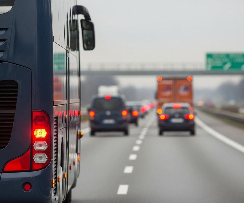 Z tyłu ciemnoniebieskiego autobusu widać włączone światła pozycyjne i kierunkowskaz po lewej stronie. Autobus stoi na asfalcie drogi z białymi przerywanymi liniami, obok pasów ruchu. Przed nim, w oddali, znajdują się niewyraźne sylwetki samochodów z czerwonymi światłami stopu, a jeszcze dalej widoczne są pomarańczowe ciężarówki i znaki drogowe na niebie.