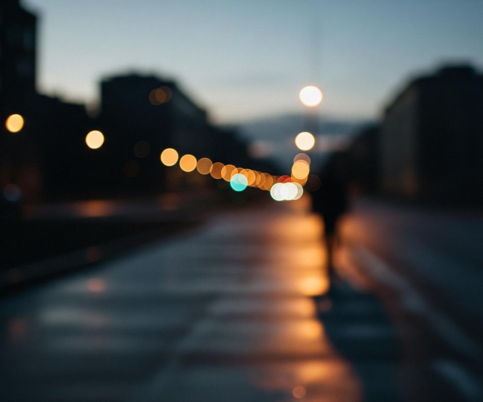 Rozmyta ulica o zmierzchu lub w nocy, z niewyraźnymi światłami latarni i sygnalizacji świetlnej tworzącymi kolorowe, okrągłe plamy. Postać ludzka, również rozmazana, widoczna jest w oddali, stojąca na chodniku i oświetlona pomarańczowym blaskiem. Architektura budynków po obu stronach drogi jest ledwo zarysowana w ciemności, a niebo ma odcień granatowy.