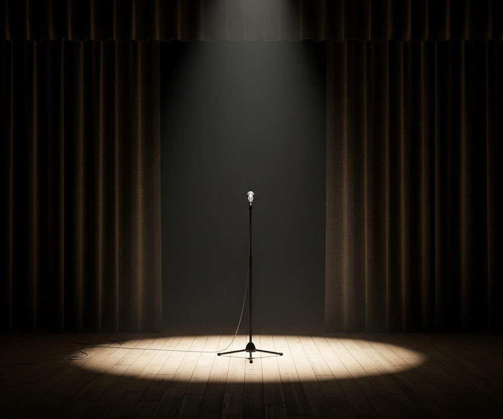 Centralnie na drewnianej scenie stoi pojedynczy, chromowany mikrofon na czarnym statywie, oświetlony mocnym, białym reflektorem z góry. Reflektor tworzy jasny okrąg światła na podłodze sceny i rozświetla otoczenie mikrofonu, podczas gdy reszta przestrzeni pozostaje w półmroku. Po obu stronach mikrofonu widoczne są ciężkie, brązowe kurtyny o pofałdowanej strukturze, otwierające się na ciemne tło. Na podłodze, po lewej stronie, z daleka od mikrofonu, leży zwinięty kabel.
