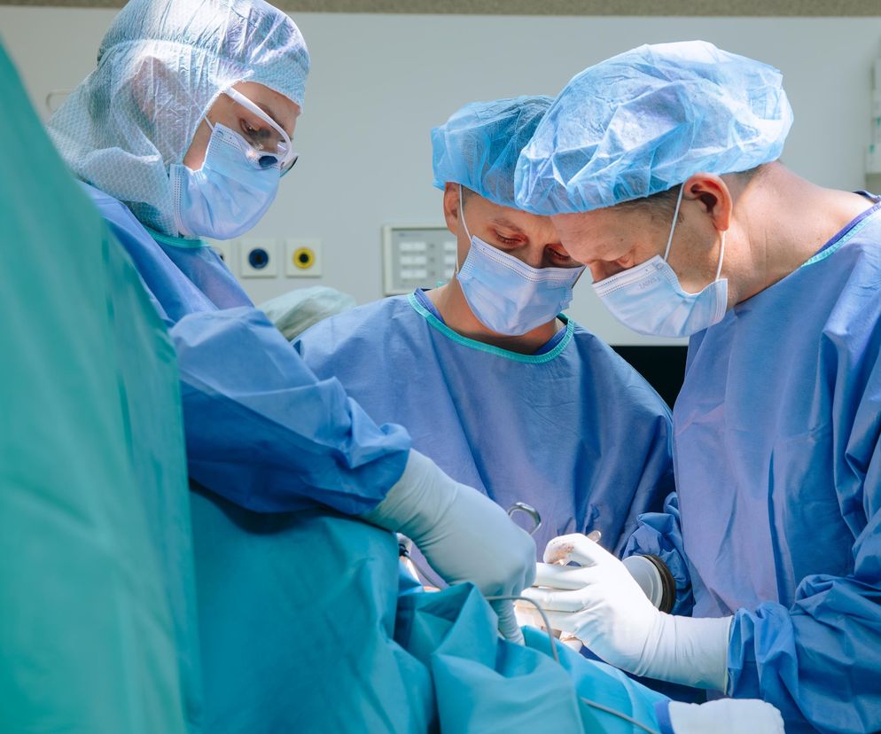 Trzej lekarze w czepkach chirurgicznych, maseczkach i sterylnych fartuchach podczas operacji. Dwóch chirurgów precyzyjnie operuje pacjenta, trzeci asystuje, odzwierciedlając wyzwania reformy wynagrodzeń w ochronie zdrowia opisywane na Super Biznes.