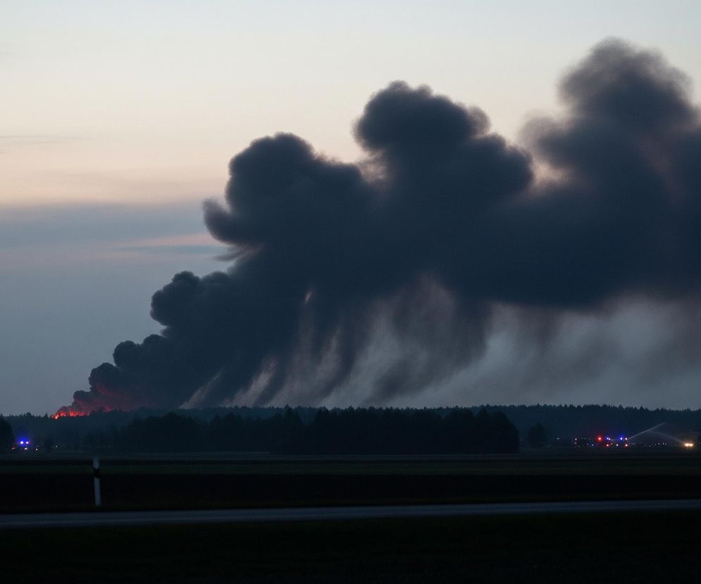 Wysokie, gęste kłęby ciemnego dymu unoszą się w górę i rozprzestrzeniają po niebie, dominując w centralnej i prawej części obrazu. U podnóża dymu, po lewej stronie, widoczne są intensywnie pomarańczowo-czerwone płomienie, zza linii ciemnych drzew, sugerujące pożar. Horyzont wyznacza pas ciemnego lasu, a przed nim rozciąga się płaski, ciemny teren, który prawdopodobnie jest polem lub łąką. Po prawej stronie, na tle lasu, widać niebieskie i czerwone migające światła, prawdopodobnie pojazdów służb ratowniczych, a także słaby strumień wody wystrzeliwujący w górę. Niebo jest jasne u góry, przechodząc w jaśniejsze odcienie szarości i błękitu ku horyzontowi.