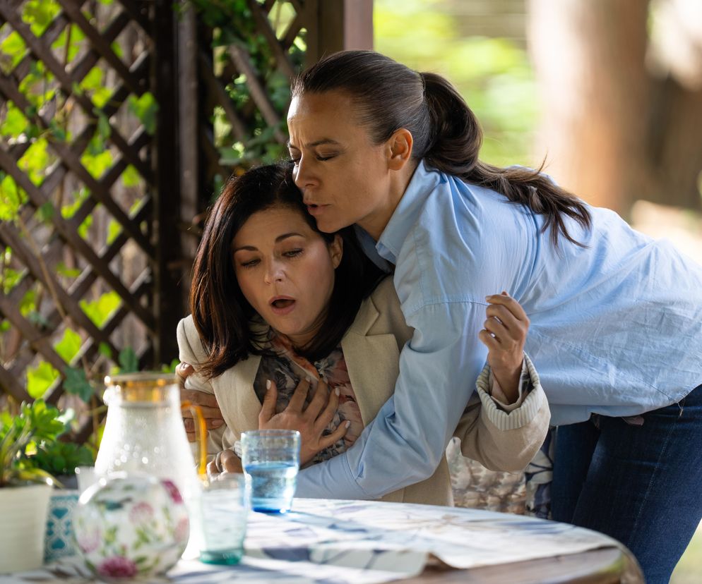 Katarzyna Cichopek (Kinga) w beżowej marynarce, z dłońmi na piersiach, walczy o oddech, wspierana przez Annę Muchę (Magdę) w błękitnej koszuli, która czule przytula ją od tyłu. Scena z M jak miłość przedstawia atak paniki Kingi po molestowaniu, na tle drewnianej altany. Więcej o tym na Super Seriale.