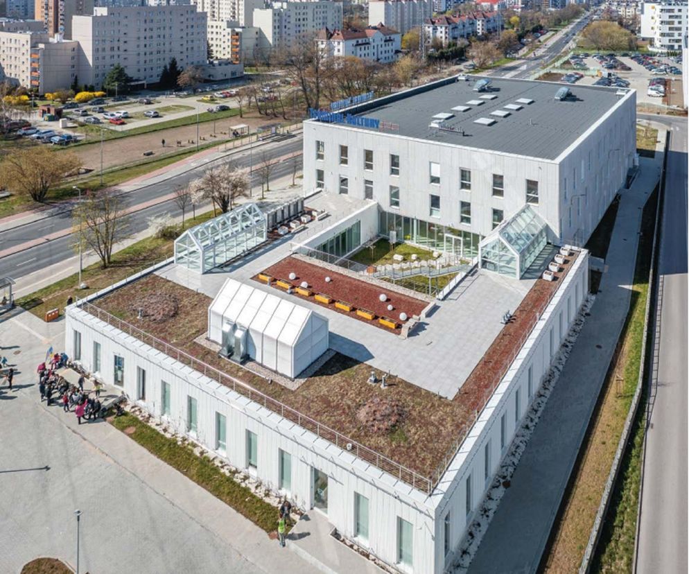 Terminal Kultury Gocław w Warszawie, widziany z lotu ptaka, prezentuje nowoczesną architekturę z zielonym dachem i przeszklonymi oranżeriami. W tle osiedle mieszkalne. Projekt autorstwa Biura 87a architekci zasługuje na uwagę, o czym przeczytasz na Architektura Murator Plus.