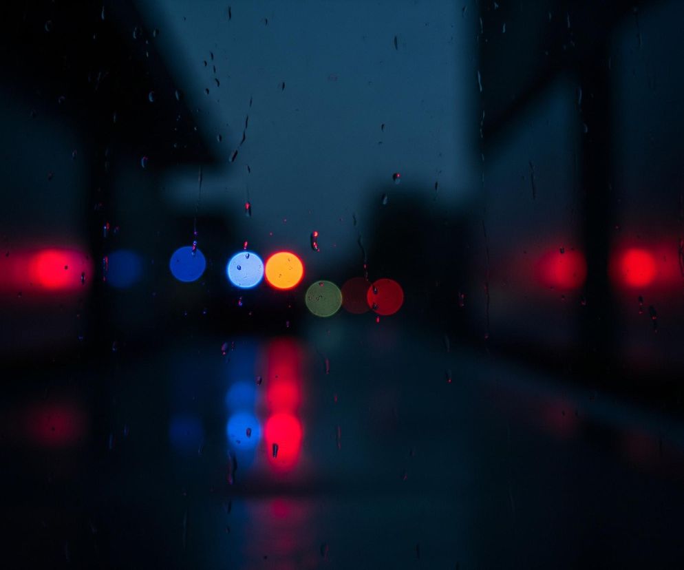 Szyba pokryta kroplami deszczu rozmywa tło ciemnej, miejskiej nocy. Na środku obrazu widoczne są rozmyte, okrągłe światła w kolorach czerwonym, niebieskim, pomarańczowym i zielonym, sugerujące ruch uliczny. Po bokach znajdują się większe, rozmazane plamy czerwonego światła, odbijające się również na dole obrazu. Dominują ciemne odcienie granatu i czerni.