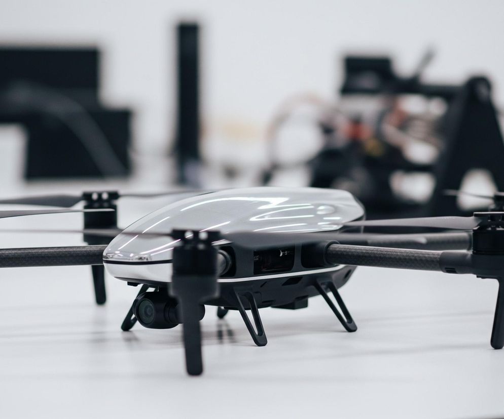 Na pierwszym planie, centralnie, leży nowoczesny dron z czterema śmigłami, skierowany lekko w prawo. Górna część jego korpusu jest lśniąco szara, z refleksami świetlnymi, natomiast dolna część i ramiona są matowo czarne. Każde ramię, wykonane z ciemnego materiału przypominającego włókno węglowe, kończy się silnikiem ze śmigłem i małym, zielonym światłem. Poniżej centralnej części korpusu widać małą, czarną kamerę z obiektywem. Dron spoczywa na białej, gładkiej powierzchni, a w tle widać rozmyte, ciemne kształty na jasnym tle, sugerujące wnętrze pomieszczenia z innymi przedmiotami.