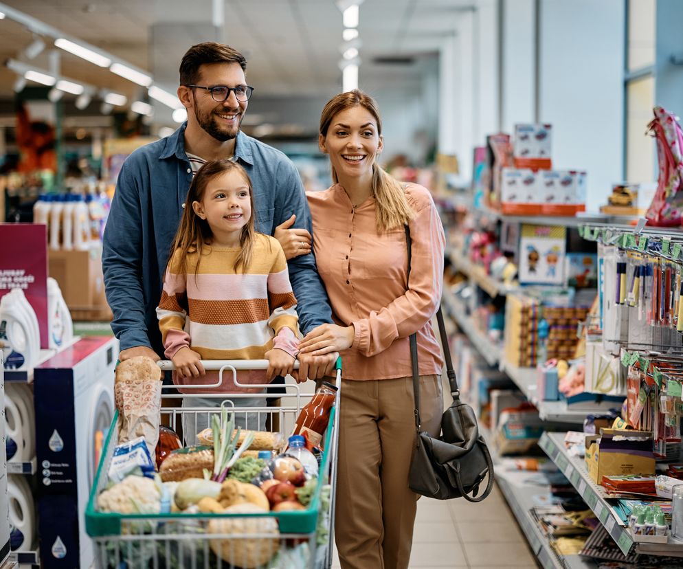 Uśmiechnięta rodzina z dzieckiem podczas zakupów w supermarkecie, pchająca koszyk pełen produktów. Obraz symbolizuje swobodę robienia zakupów w dni handlowe, o których dowiesz się na Super Biznes.
