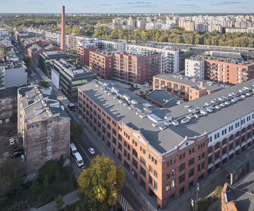 Widok z lotu ptaka na zrewitalizowany kompleks Bohema Praga przy ul. Szwedzkiej w Warszawie, ukazujący ceglane budynki mieszkalne i usługowe z elementami poprzemysłowymi oraz dominujący komin, harmonizujące z otoczeniem i historyczną zabudową. Więcej na Architektura Murator Plus.