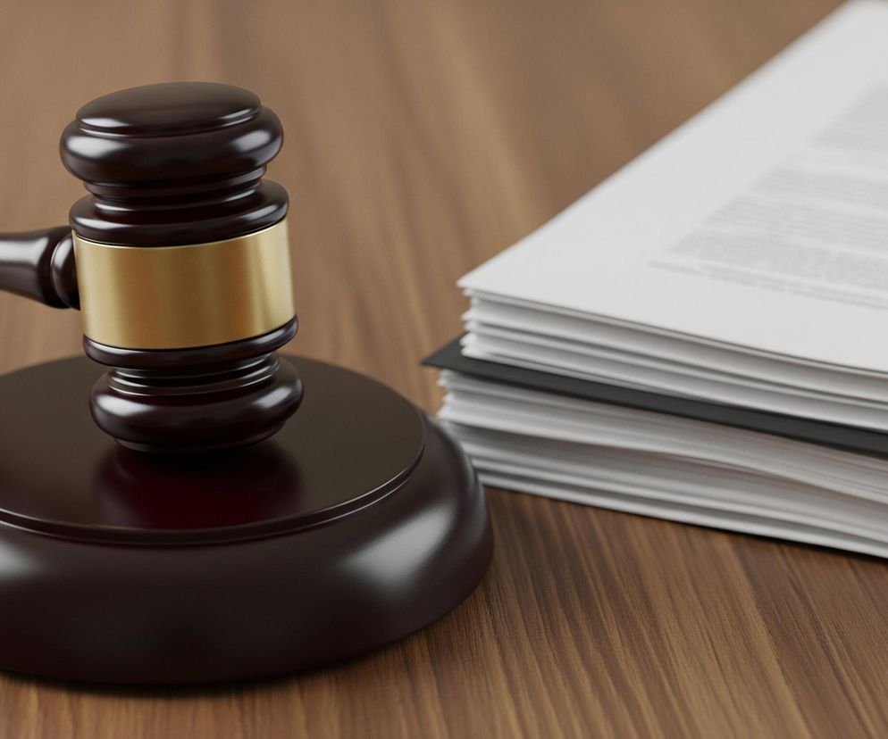 Na drewnianym, brązowym blacie stołu leży ciemnobrązowy młotek sędziowski z okrągłą, złotą wstawką na główce, umieszczony na dopasowanej, okrągłej podstawie. Po prawej stronie, w tle, widoczny jest stos białych, zadrukowanych kartek, częściowo zakryty cieniem. Obraz przedstawia scenę z perspektywy lekko z góry, z ostrością na młotku, podczas gdy stos dokumentów jest nieznacznie rozmyty.