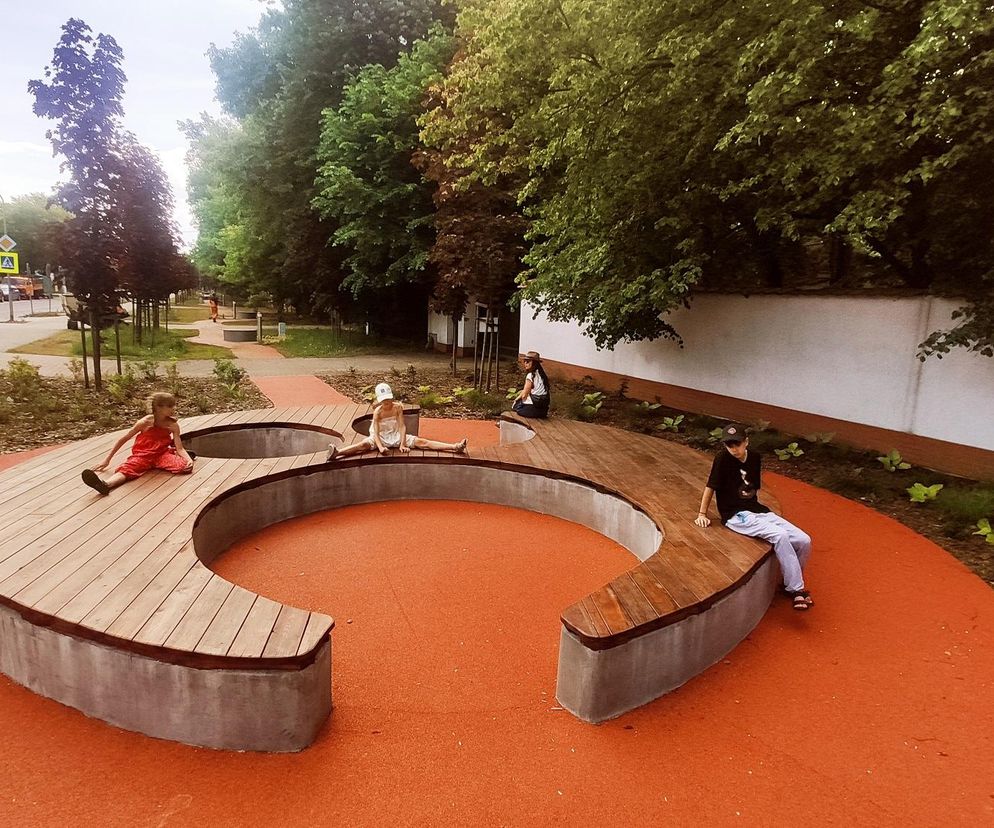 Park Artystów w Łomiankach, zaprojektowany przez Gdak Studio, z pomarańczową, wijącą się ścieżką i drewnianymi, zaokrąglonymi ławkami, na których siedzą dzieci. To projekt zgłoszony do konkursu ŻYCIE W ARCHITEKTURZE 2025, o którym przeczytasz na Architektura Murator Plus.
