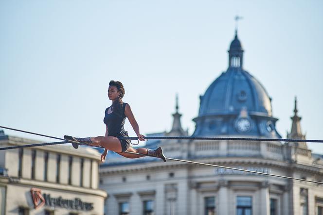 Kobieta wykonuje szpagat na rozpiętej w powietrzu linie. W tle dach Grand Hotelu w Lublinie