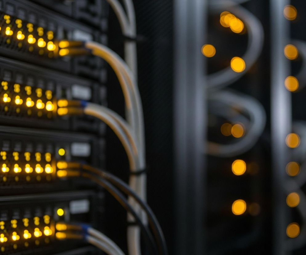 Cztery czarne, prostokątne urządzenia elektroniczne, ułożone jeden nad drugim w rzędzie, wypełniają lewą stronę kadru. Każde z urządzeń posiada szereg małych, jasnożółtych świecących lampek, zlokalizowanych po prawej stronie od głównego włącznika. Z tych urządzeń wychodzą kable w kolorze szarym, z niebieskimi końcówkami, biegnące w prawą stronę. Tło obrazu jest ciemne i rozmyte, z widocznymi, jasnymi, żółtymi plamami światła, tworzącymi efekt bokeh.