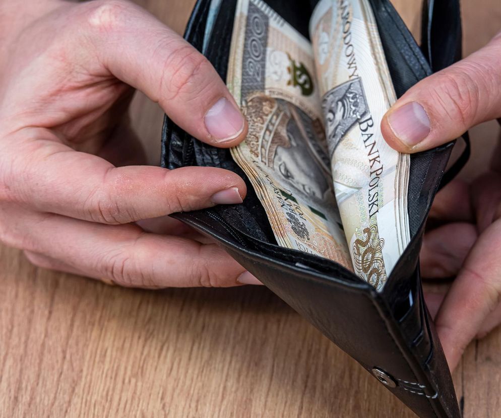 Dwie dłonie otwierają czarny portfel, z którego wystają złożone banknoty o nominale 200 złotych, ułożone na jasnym drewnianym tle. Zdjęcie symbolizuje kwestię finansowania świadczeń dla niepełnosprawnych, o której przeczytasz na Super Biznes.