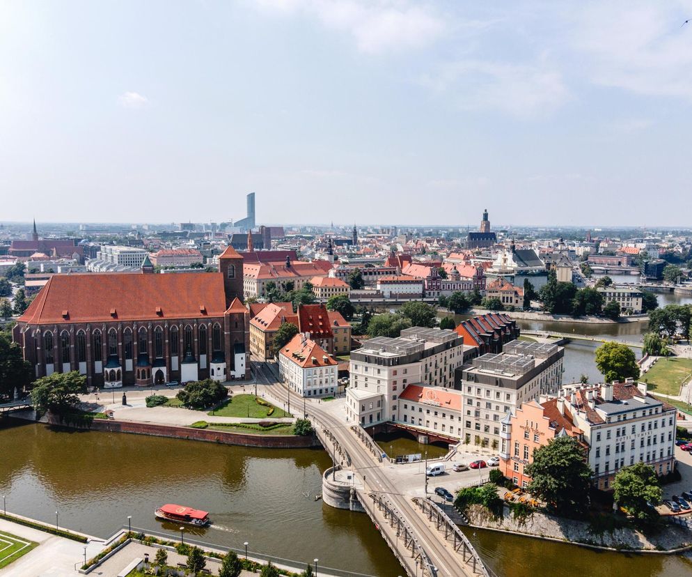 Panorama Wrocławia z góry, prezentująca Ostrów Tumski z Katedrą św. Jana Chrzciciela i okolicznymi budynkami mieszkalnymi oraz Most Tumski nad Odrą. Widać rzekę z pływającą łodzią wycieczkową oraz dachy kamienic. Więcej o architekturze Wrocławia przeczytasz na Architektura Murator Plus.