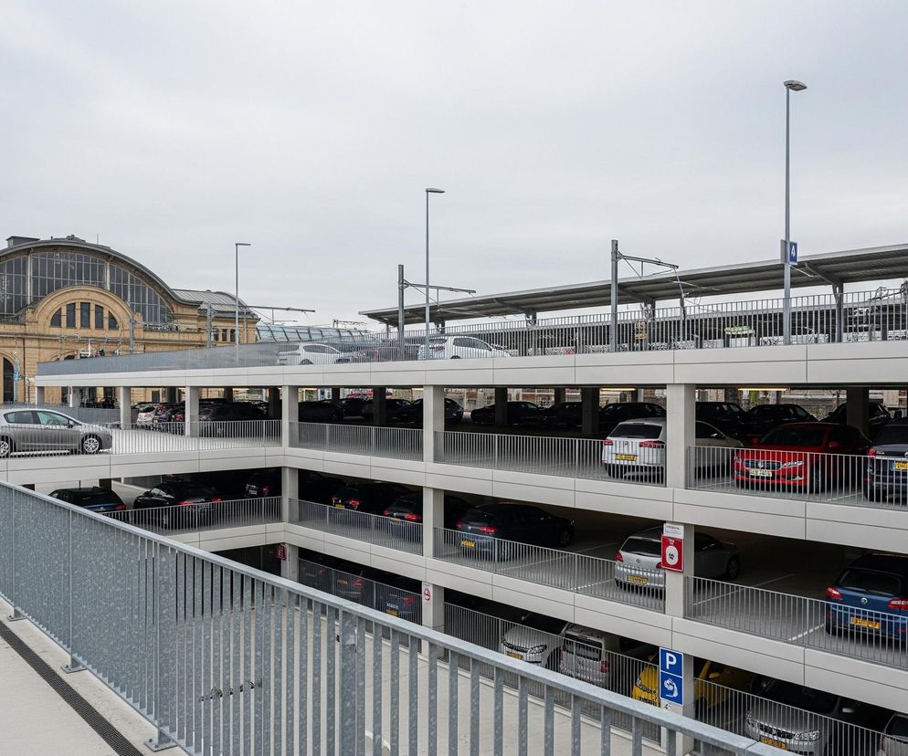 Duży, wielopoziomowy parking dla samochodów zajmuje centralną i prawą część obrazu, z fasadą zabytkowego budynku w tle po lewej stronie. Na parkingu stoją liczne samochody różnych marek i kolorów, zaparkowane na kolejnych piętrach, widocznych przez metalowe barierki. Nad najwyższym poziomem parkingu znajduje się szary, zadaszony obszar, a na jego tle widać latarnie uliczne i słupy z liniami energetycznymi. W dolnej lewej części obrazu znajduje się betonowy chodnik z metalową barierką, która biegnie przez całą długość kadru.