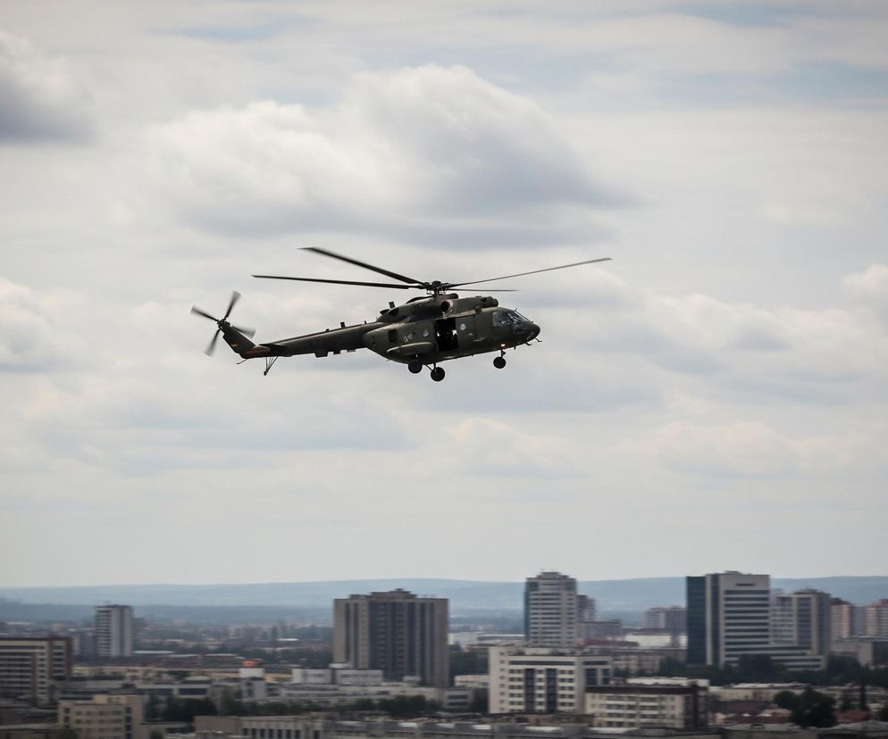 Duży wojskowy helikopter w kolorze ciemnooliwkowym, z wyraźnie widocznymi obracającymi się łopatami wirnika głównego i ogonowego, przelatuje na tle częściowo zachmurzonego nieba. Pod nim rozciąga się panorama miasta z licznymi wysokimi budynkami, a w oddali widać zarys pagórków lub pasma górskiego. Całość sprawia wrażenie szerokiego ujęcia, przedstawiającego helikopter w locie nad miejskim krajobrazem.
