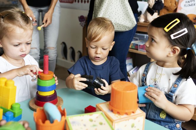 Troje dzieci w wieku przedszkolnym siedzi przy stoliku i bawi się różnokolorowymi zabawkami edukacyjnymi. Dziewczynka w białej koszulce z biżuterią układa piramidę z klocków, obok chłopiec w granatowej koszulce składa czarne elementy. Przy nim dziewczynka w ogrodniczkach i białej bluzce. Tematy dotyczące zdrowia dzieci można przeczytać na portalu Poradnik Zdrowie.