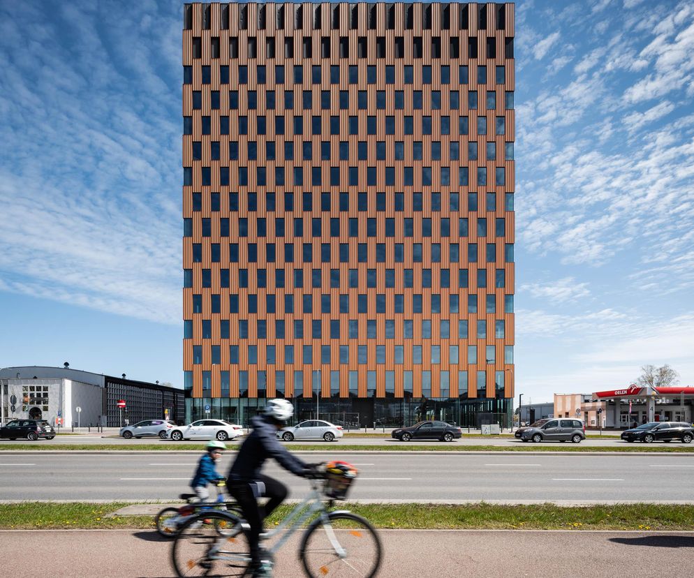 Nowoczesny budynek biurowy Wave w Gdańsku, autorstwa Medusa Group, z bursztynową elewacją z pionowymi podziałami. Na pierwszym planie rozmazana sylwetka rowerzysty na tle ruchliwej ulicy. Projekt nominowany w konkursie Życie w Architekturze, o czym można przeczytać na Architektura Murator Plus.