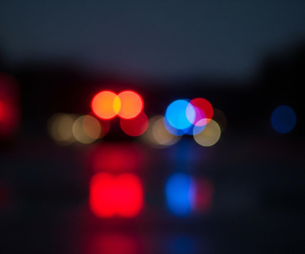 Rozmyte światła na ciemnym tle dominują obraz. W centrum widoczne są dwa duże, okrągłe światła w kolorze czerwono-pomarańczowym, częściowo nałożone na siebie, obok nich po prawej stronie znajduje się niebieskie, okrągłe światło oraz kilka mniejszych, jaśniejszych, okrągłych punktów świetlnych w odcieniach czerwieni i beżu. Poniżej tych świateł widoczne są ich rozmyte odbicia w intensywnych barwach czerwieni i błękitu, sugerujące mokrą powierzchnię. Tło jest ciemnogranatowe, niemal czarne, z delikatnym gradientem ku górze, gdzie wydaje się jaśniejsze.