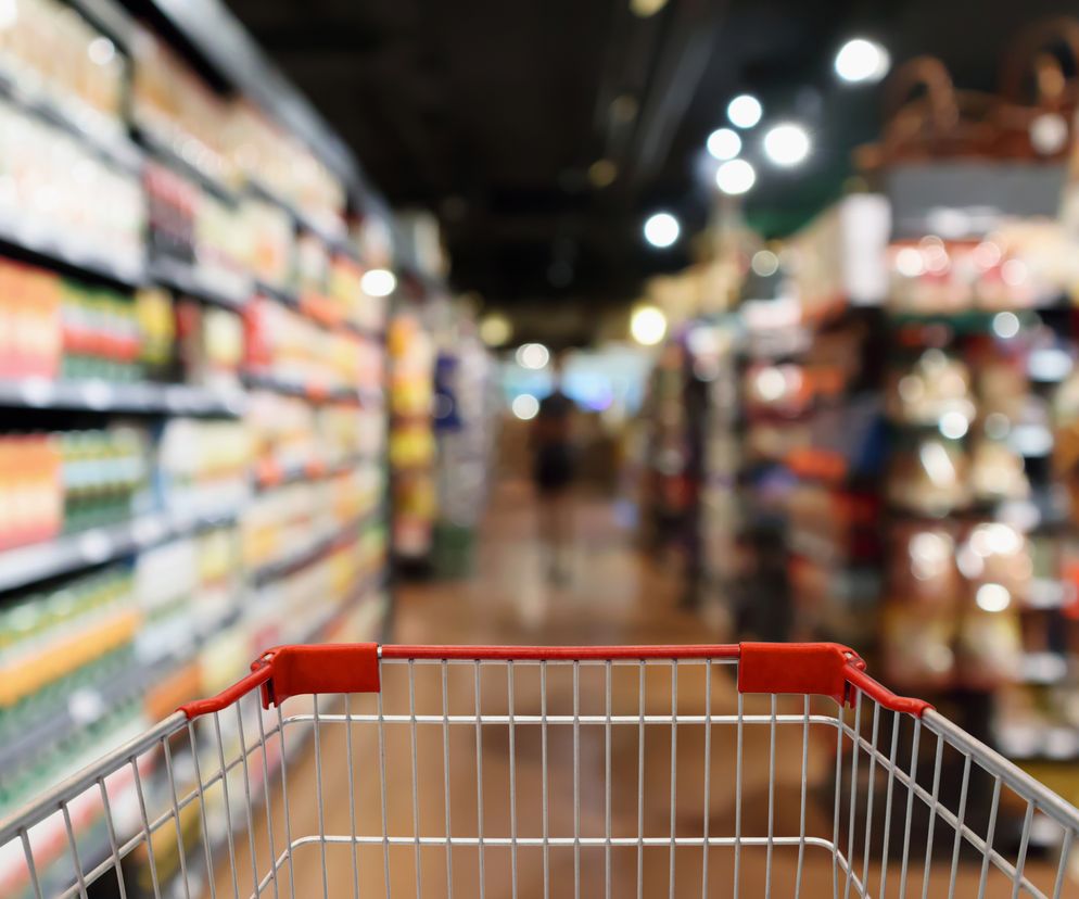Pusty wózek sklepowy na pierwszym planie, z czerwonym uchwytem i chromowanymi elementami. W tle rozmazany widok alejek z półkami pełnymi produktów w supermarkecie. Zdjęcie ilustruje temat ponownego otwierania sklepów i galerii handlowych, o czym można przeczytać na portalu Poradnik Zdrowie.