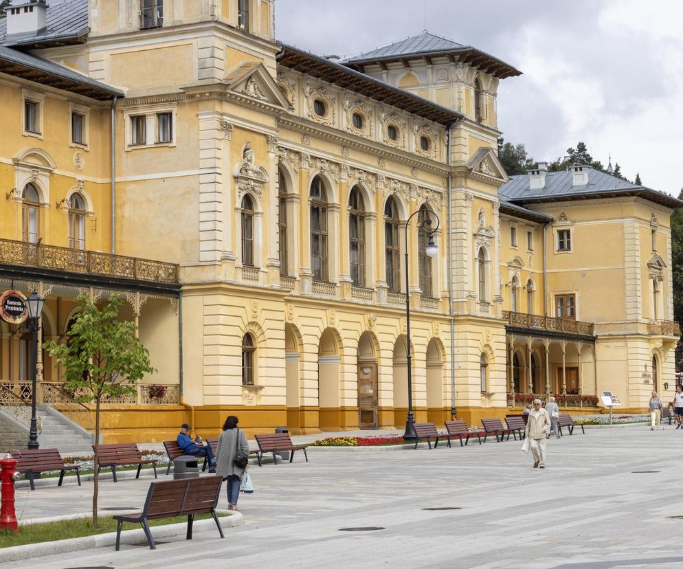 Elegancki budynek uzdrowiskowy z jasną fasadą i ozdobnymi detalami, widoczny od frontu z dużym placem, ławkami i grupą spacerujących osób. To idealny przykład architektury uzdrowiskowej, o której można przeczytać na portalu Poradnik Zdrowie.