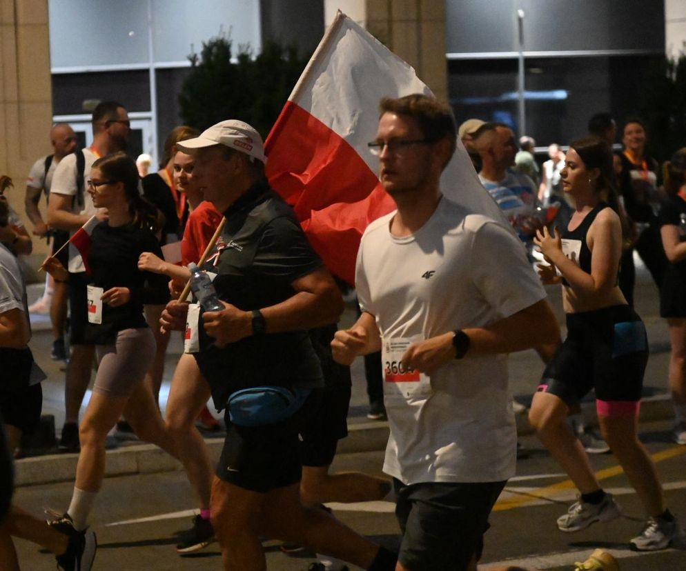 Grupa osób biegnąca ulicą po zmroku. Jeden z mężczyzn trzyma polską flagę. O Biegu Powstania Warszawskiego przeczytasz na SE.