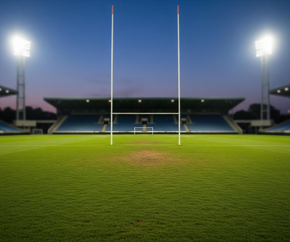 Boisko pokryte zieloną trawą rozciąga się w kierunku widowni i nieba. Centralnie na boisku stoi bramka do rugby w kształcie litery H, z dwoma wysokimi białymi słupkami połączonymi poziomą poprzeczką. Dwa słupki mają czerwone akcenty na szczytach. W tle widać rzędy pustych, niebieskich siedzeń stadionu, a ponad nimi ciemne niebo z lekkim fioletowym gradientem. Po lewej i prawej stronie stadionu stoją jasne reflektory, oświetlające całe boisko.