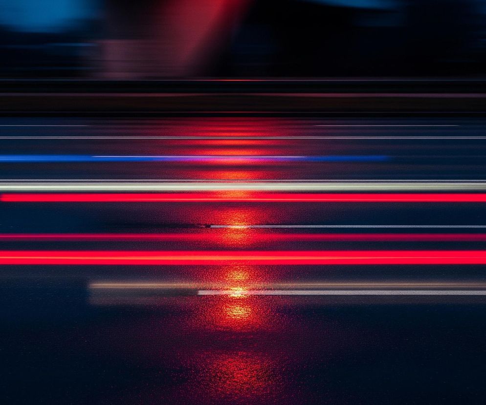 Ciemną, poziomą drogę nocą, z rozmytymi smugami świateł pojazdów tworzącymi dynamiczne linie. Na pierwszym planie widać szeroki, czarny pas asfaltu z widoczną teksturą, na którym odbijają się intensywne czerwone, białe i niebieskie pasma światła, rozciągające się horyzontalnie przez całą szerokość kadru. Górna część obrazu to ciemniejsze, rozmyte tło, gdzie również dostrzegalne są niewyraźne, poziome linie, sugerujące budynki lub infrastrukturę miejską. Dominują kolory ciemnego granatu, czerni i intensywnych barw czerwieni, bieli oraz błękitu.