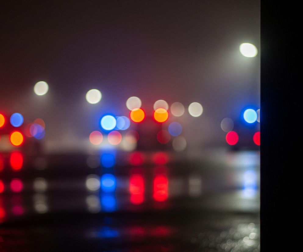 Na pierwszym planie widoczna jest rozmyta scena nocna z ulicznymi światłami w postaci jasnych, okrągłych plam bokeh. Przeważają kolory czerwony i niebieski, co sugeruje światła pojazdów uprzywilejowanych, oraz żółtawe i białe plamy pochodzące od latarni lub innych źródeł światła. Powierzchnia drogi jest mokra, odbijając kolorowe światła i tworząc podłużne, rozmyte refleksy. Prawa krawędź obrazu jest ciemna, z pionową czarną powierzchnią zasłaniającą część sceny.