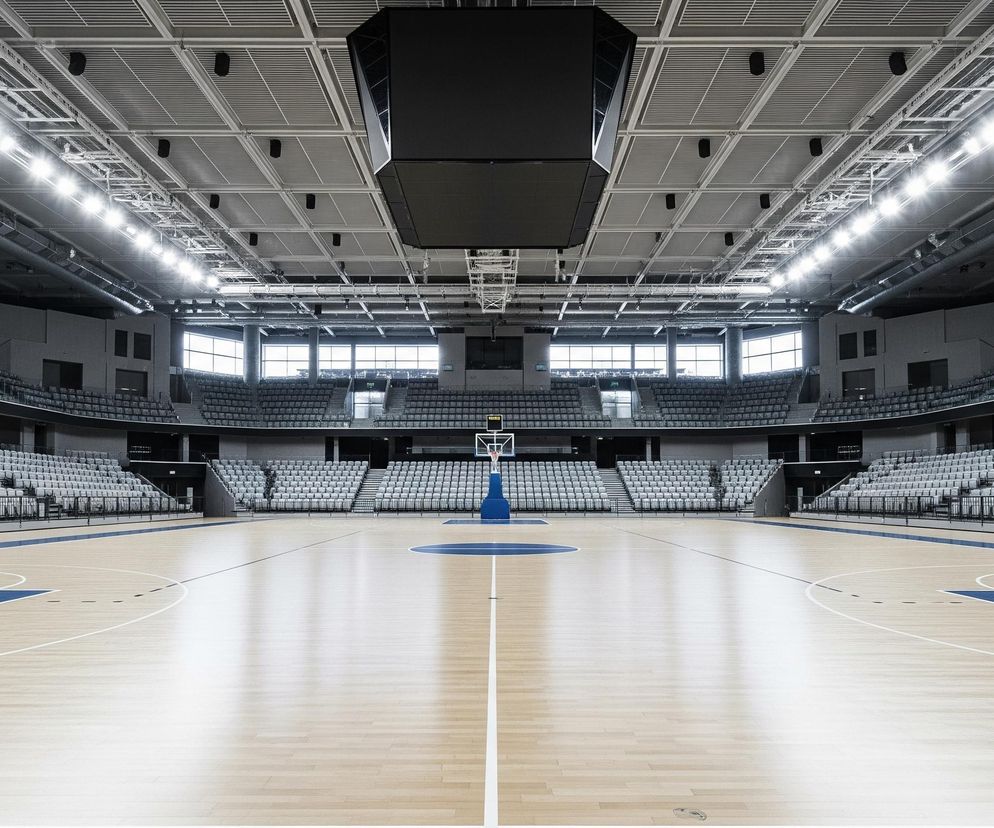 Wnętrze nowoczesnej hali sportowej z drewnianą, lśniącą podłogą boiska do koszykówki, oznaczoną białymi liniami i niebieskimi obszarami. Na środku stoi niebieska konstrukcja kosza z białą obręczą i siatką. Wokół boiska rozciągają się puste trybuny z szarymi siedzeniami, rozmieszczone w kilku poziomach, a powyżej nich widoczne są duże okna. Sufit jest jasny, kratownicowy, z rzędami jaskrawo świecących lamp oraz zawieszonym centralnie, dużym, czarnym telebimem.
