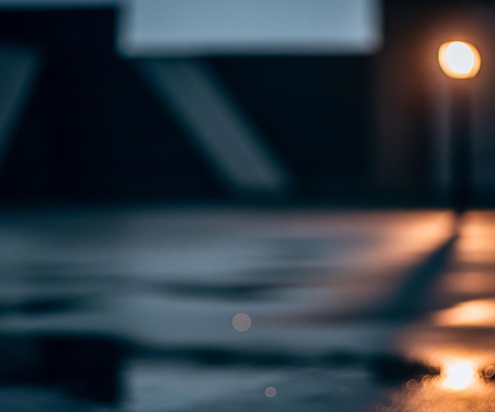 Jasna, pomarańczowo-biała kula światła znajduje się po prawej stronie zdjęcia, tworząc pionową smużkę rozciągającą się w dół do dołu kadru. To światło odbija się w ciemnej, wilgotnej powierzchni, która pokrywa cały dolny obszar obrazu. Górna część zdjęcia jest ciemna, z rozmytymi, jaśniejszymi szaro-niebieskimi elementami architektonicznymi po lewej stronie, zlewającymi się z tłem.