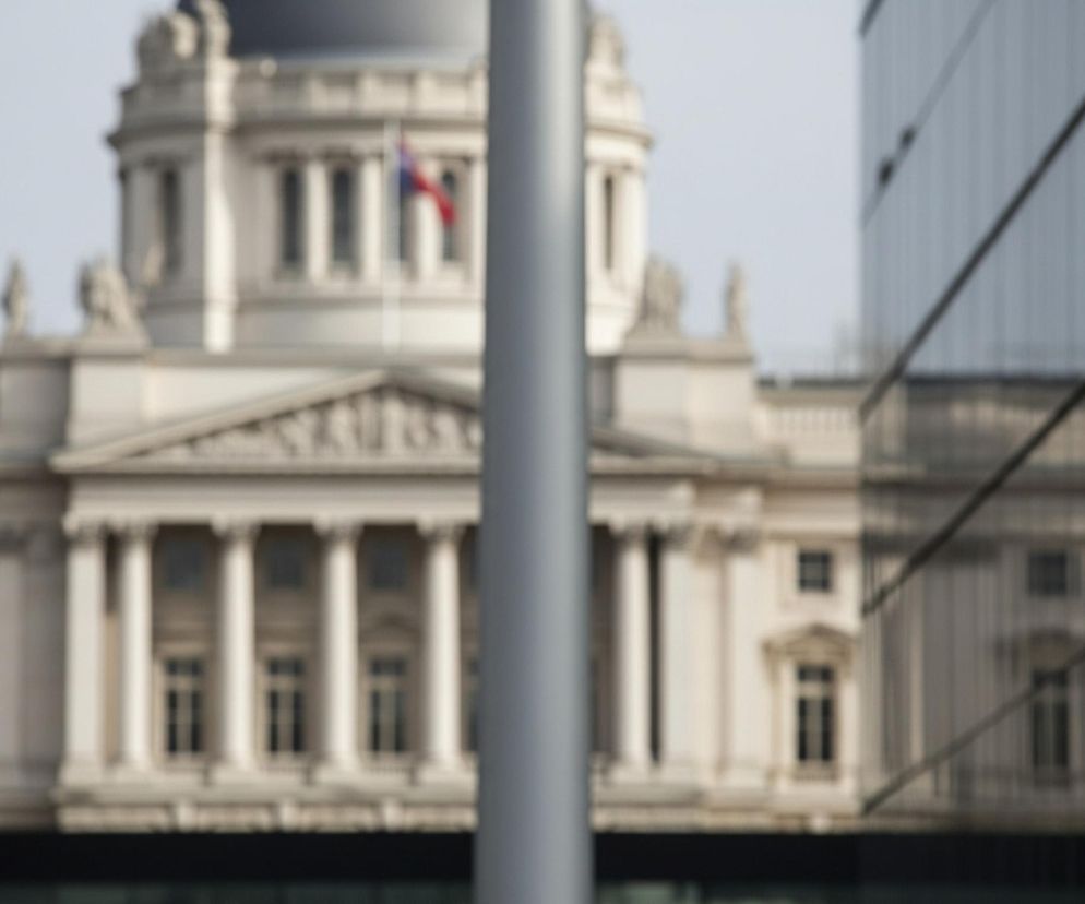 Centralnie na zdjęciu znajduje się szare, niewyraźne słup o pionowej orientacji, rozciągający się od góry do dołu. Za nim, w tle, widoczny jest rozmyty, jasny budynek z kopułą, ozdobiony kolumnami i okapnikami, z flagą o czerwono-niebieskich barwach powiewającą na masztu. Po prawej stronie słupa, również rozmyty, widnieje fragment ciemnego, lustrzanego budynku z poziomymi i ukośnymi liniami odbicia. Całe zdjęcie ma płytką głębię ostrości, przez co większość elementów jest nieostra.