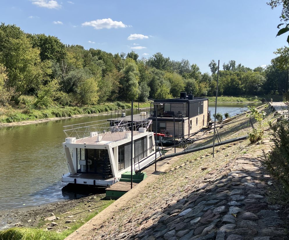 Dwa nowoczesne domy na wodzie zacumowane przy kamienistym brzegu rzeki. O houseboatach w Polsce przeczytasz na Muratordom.