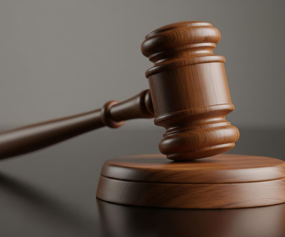 Młotek sędziowski wykonany z drewna, o wyraźnym usłojeniu w odcieniach brązu. Składa się z rękojeści i obuchu o cylindrycznym kształcie, z pogrubionym, żłobionym fragmentem. Młotek leży na okrągłej drewnianej podstawie, również o brązowym zabarwieniu i widocznym usłojeniu, a tło stanowi gładka, jednolita szarość, częściowo rozjaśniona u góry. Całość jest oświetlona z góry, tworząc refleksy i cienie na powierzchniach.