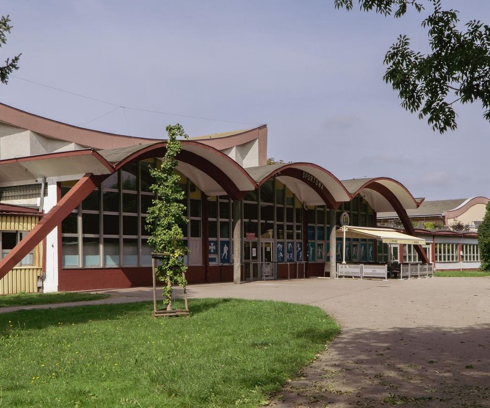 Hala sportowa ROSiR w Rzeszowie, z charakterystycznymi łukowatymi zadaszeniami wejścia. Budynek otoczony jest zielenią i fragmentami chodnika. O jej niezwykłej architekturze przeczytasz na Architektura Murator Plus.