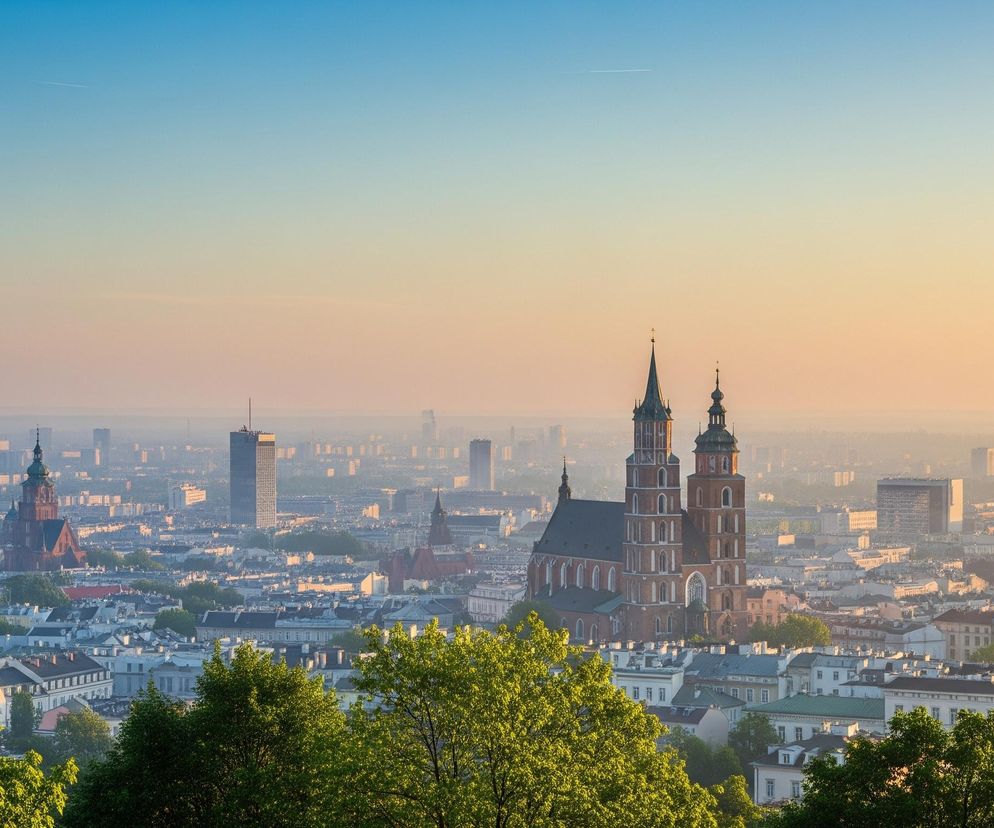 Centralnym elementem obrazu jest duży, ceglany kościół z dwiema wieżami o różnej wysokości, zlokalizowany w centrum panoramy miasta. Dachy kościoła są ciemnoszare, a ściany i wieże mają intensywny, ceglasty kolor. W tle rozciąga się miejski krajobraz z licznymi budynkami mieszkalnymi o jasnych elewacjach oraz kilkoma wysokimi drapaczami chmur. Całość jest skąpana w delikatnym, złotym świetle poranka lub wieczoru, które rozmywa odległe detale. Na pierwszym planie widoczne są intensywnie zielone korony drzew, częściowo zasłaniające zabudowania, a niebo stopniowo przechodzi od jasnego błękitu u góry do cieplego, pomarańczowego odcienia tuż nad horyzontem.