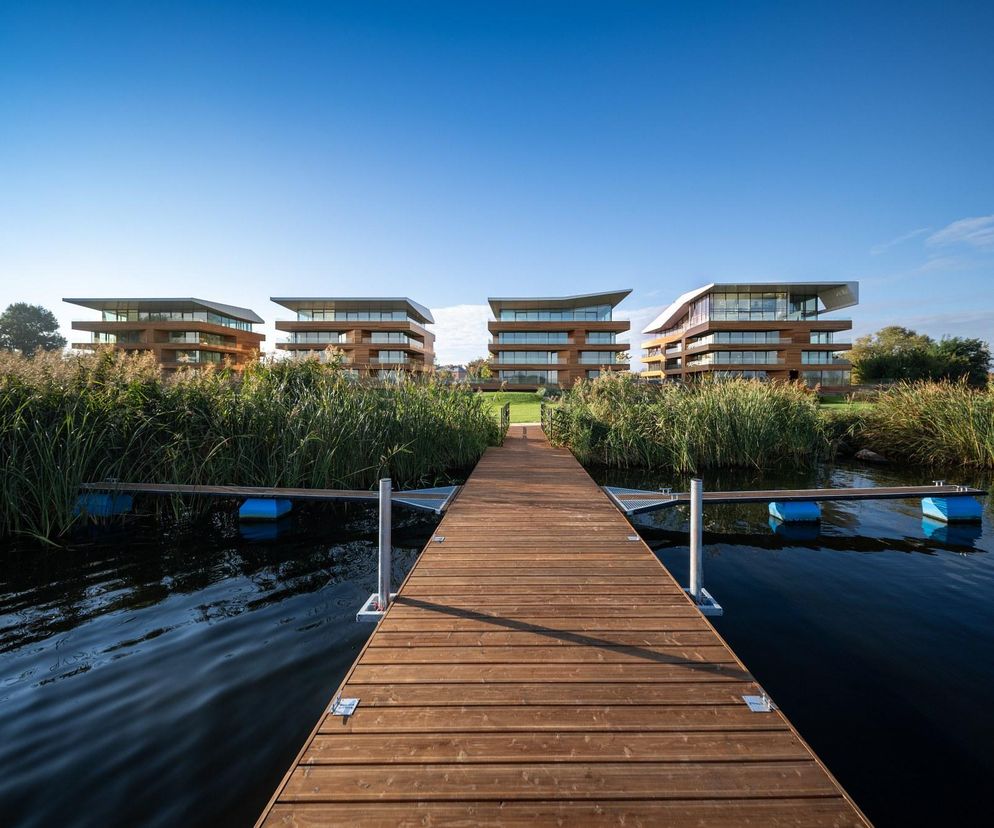 Drewniany pomost prowadzący przez szuwary do kompleksu nowoczesnych budynków apartamentowych z funkcją turystyczną Riva Zegrze. Obiekty o drewnianej elewacji i płaskich dachach harmonijnie wpisują się w krajobraz Jeziora Zegrzyńskiego, o czym więcej przeczytasz na Architektura Murator Plus.