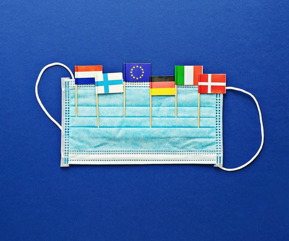 Niebieska maseczka ochronna leżąca na ciemnoniebieskim tle, z patyczkami z flagami różnych krajów europejskich symbolizującymi znoszenie ograniczeń pandemicznych. Flagi: Holandii, Finlandii, Unii Europejskiej, Niemiec, Włoch i Danii. Więcej o znoszeniu obostrzeń na Poradnik Zdrowie.