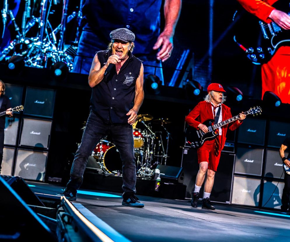 Zespół AC/DC podczas energicznego występu na scenie. Brian Johnson w czarnej koszulce i kaszkiecie śpiewa do mikrofonu, a Angus Young w charakterystycznym czerwonym szkolnym mundurku gra na gitarze elektrycznej, obok pozostali muzycy. O tym, ile zarobiły zespoły rockowe na koncertach, przeczytasz na naszym portalu.