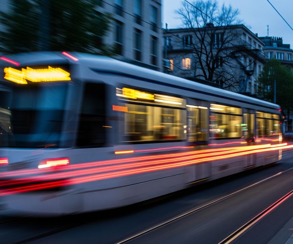 Biały tramwaj z rozświetlonymi żółtymi oknami i czerwonymi światłami bocznymi pędzi po torach, pozostawiając za sobą długie, czerwone smugi świateł pozycyjnych na tle ciemniejącego nieba. Obok niego biegnie ulica z ruchem ulicznym, gdzie widać jasne smugi świateł samochodów. Po lewej stronie widoczne są rozmazane fasady budynków, a po prawej chodnik i drzewa.