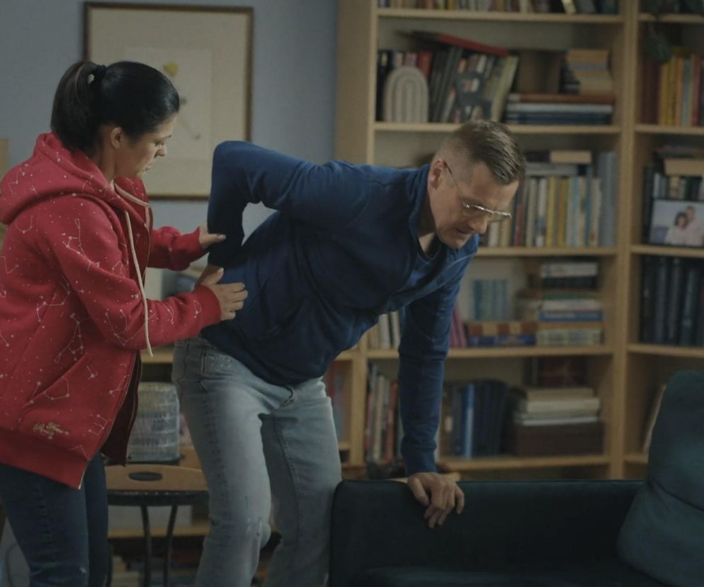 Katarzyna Cichopek jako Kinga w czerwonej bluzie ze wzorem konstelacji, wspiera Marcina Mroczka jako Piotrka w niebieskiej bluzie, który cierpi na ból pleców po wypadku w domu. Scena ukazuje panikę Kingi i ból Piotrka w 1882 odcinku M jak miłość. Więcej o serialach na Super Seriale.