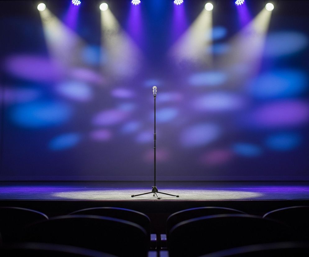 Na scenie stoi samotny mikrofon na czarnym statywie z trójnogiem, oświetlony od góry kilkoma snopami światła. Scena jest podświetlona na fioletowo-niebiesko, z jaśniejszymi plamami światła na tle. Po bokach sceny wiszą bordowe, pofałdowane zasłony. Z pierwszego planu widoczne są czarne, niewyraźne oparcia siedzeń widowni, wskazujące na pustą salę.