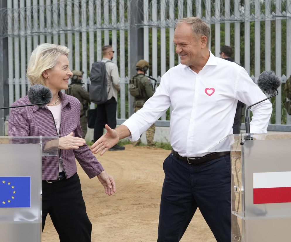 Ursula von der Leyen i Donald Tusk uśmiechają się, podając sobie ręce na tle ogrodzenia.  Na obrazku widoczne są także osoby w mundurach.  Więcej na temat nowego systemu ETS-2 oraz jego wpływu na Polskę przeczytasz na portalu Super Biznes.