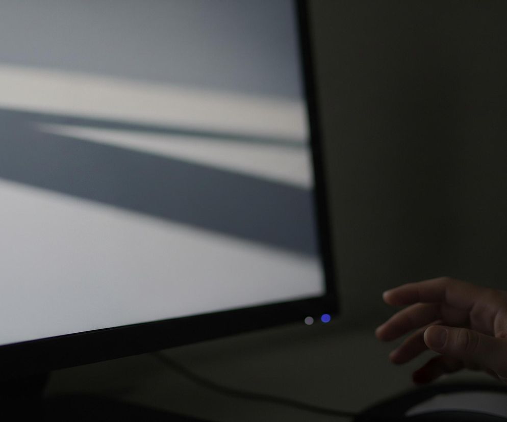 Główny plan zajmuje ekran monitora o jasnej, szarej powierzchni, na której widoczne są cienie tworzące geometryczne kształty. Lewa część ekranu jest jaśniejsza, a prawy dolny róg monitora ukazuje dwa okrągłe, świecące punkty – jeden niebieski i jeden jaśniejszy, niemal biały. Po prawej stronie kadru, na ciemnym tle, widać prawą dłoń wyciągniętą w kierunku monitora, a niżej częściowo uchwyconą mysz komputerową.