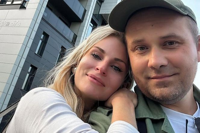 Uśmiechnięta para, Adrian Szymaniak w zielonej czapce i kobieta o blond włosach, pozuje do selfie na tle nowoczesnego budynku mieszkalnego. Zdjęcie ilustruje wyzwania związane z chorobą, o czym można przeczytać na portalu Poradnik Zdrowie.