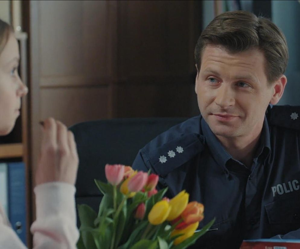 Hania i Adam Karski, komendant policji, rozmawiają w biurze. Hania z bukietem tulipanów rozmawia z Adamem w mundurze z napisem POLICJA. Scena z serialu M jak miłość w 1886 odcinku, o której można przeczytać więcej na Super Seriale.