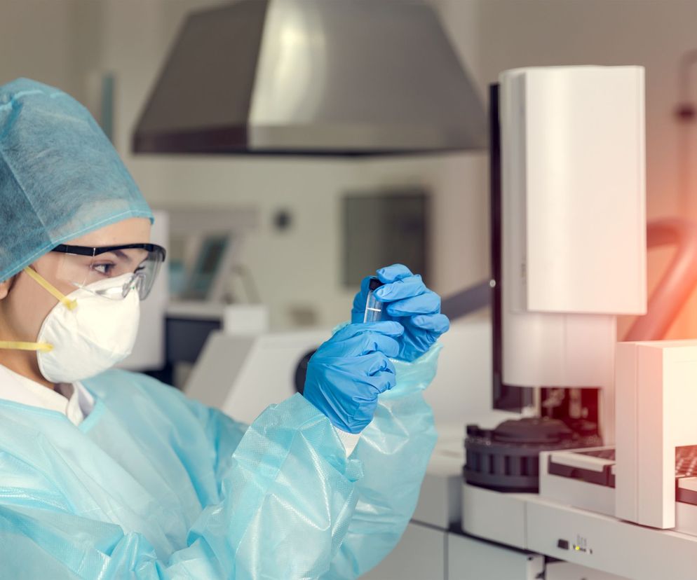 Osoba w sterylnym stroju ochronnym, masce N95, czepku i okularach laboratoryjnych, trzymająca próbówkę, przygotowuje się do analizy. Obok widoczny sprzęt diagnostyczny. Zdjęcie symbolizuje szybkie wykrywanie COVID-19, o którym przeczytasz na portalu Poradnik Zdrowie.
